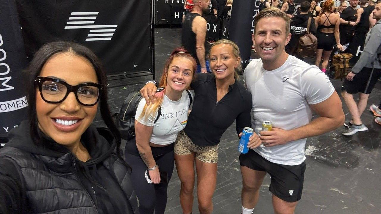 A selfie of four people who in wearing gym clothing and  standing in an indoor arena that is set up for a fitness competition. There are three women and one man. 