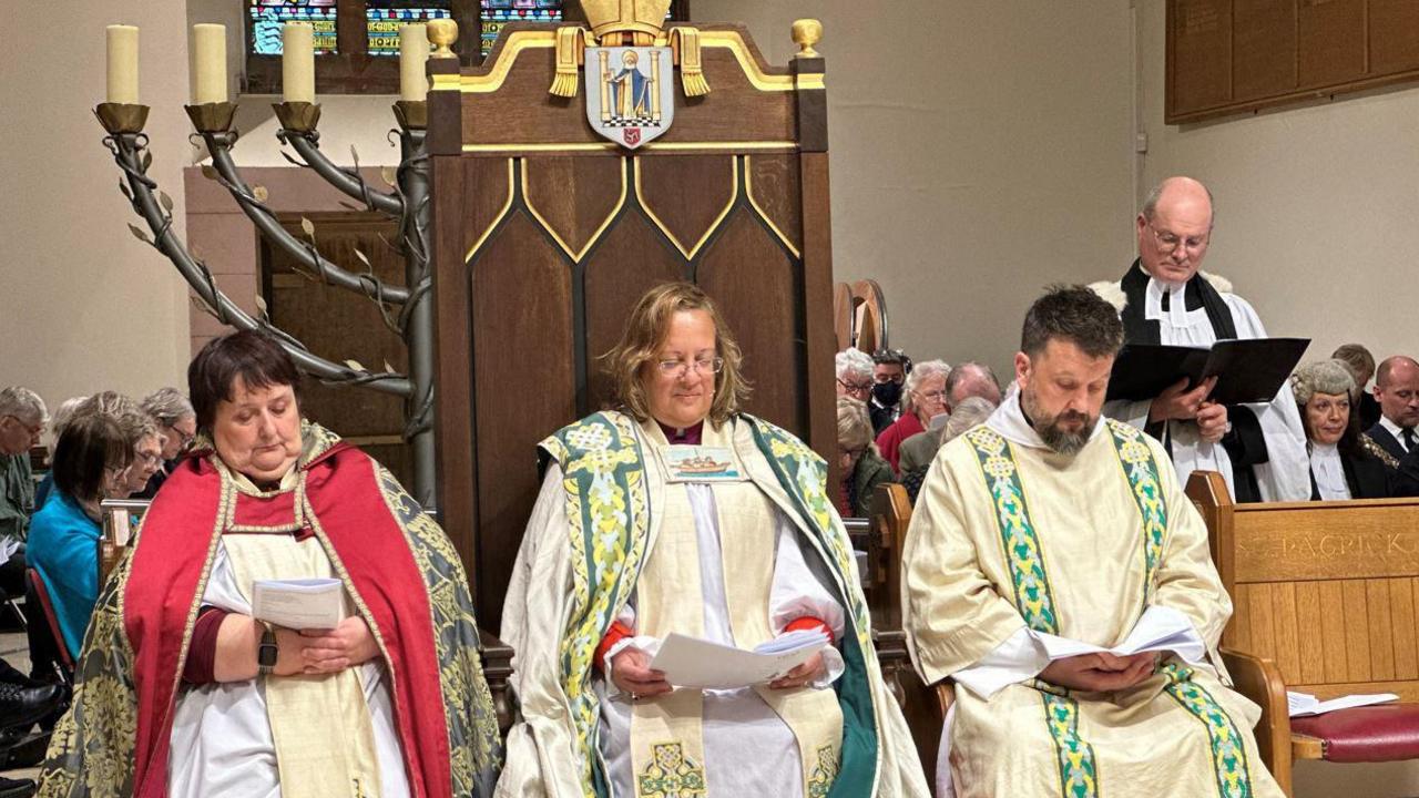 Rt Rev Tricia Hillas sitting on the bishop's throne between two other members of the clergy at a service at Cathedral Isle of Man in Peel. All three are wearing Anglican vestments. 