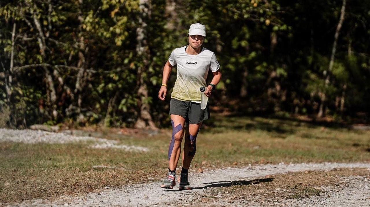 A young woman wearing a white t-shirt and black shorts and holding a water bottle runs along a dirt road. She had purple tape strapped around her knees.