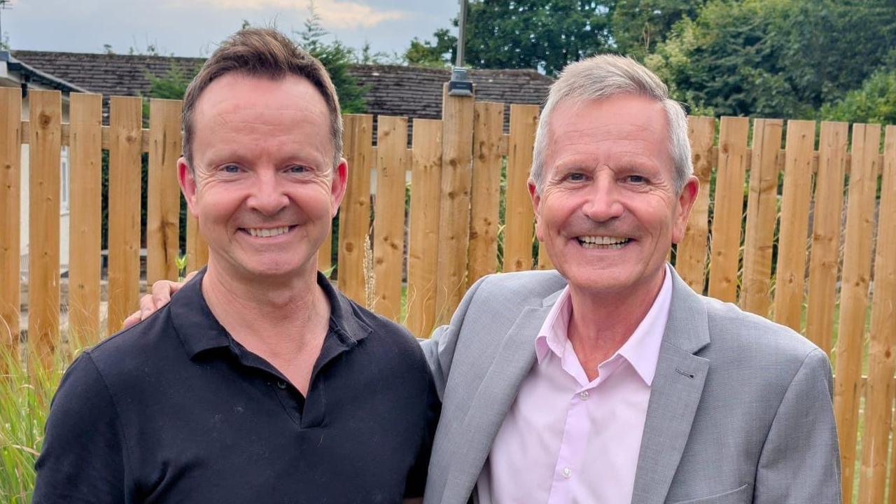 Man with short brown hair and wearing a dark shirt, standing next to another man with grey hair, who is wearing a pink shirt and grey jacket. The pair are stood in a garden with a wooden-panelled fence in the background.