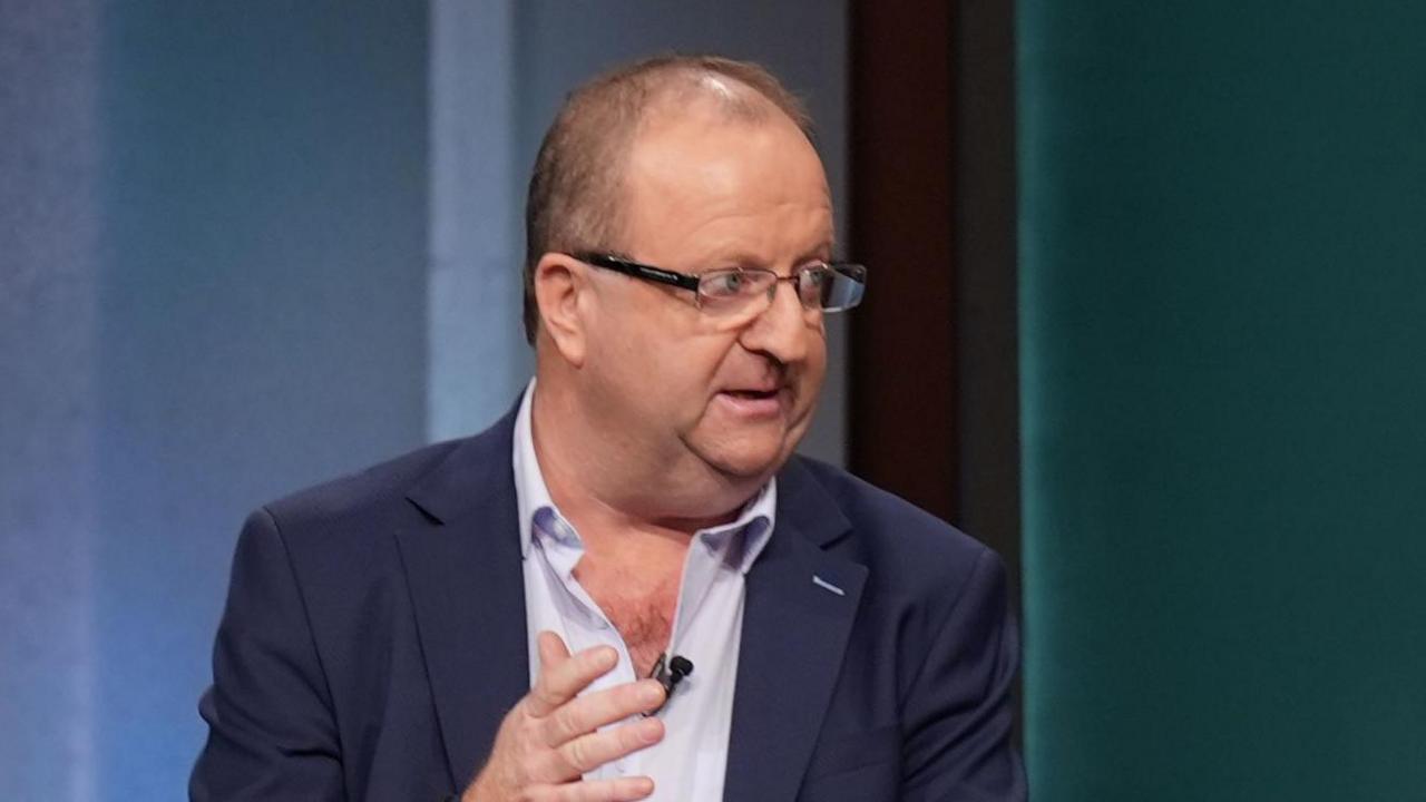 Leader of Independent Ireland, Michael Collins is bald, wearing glasses, a dark blue blazer and light blue shirt with top buttons open.