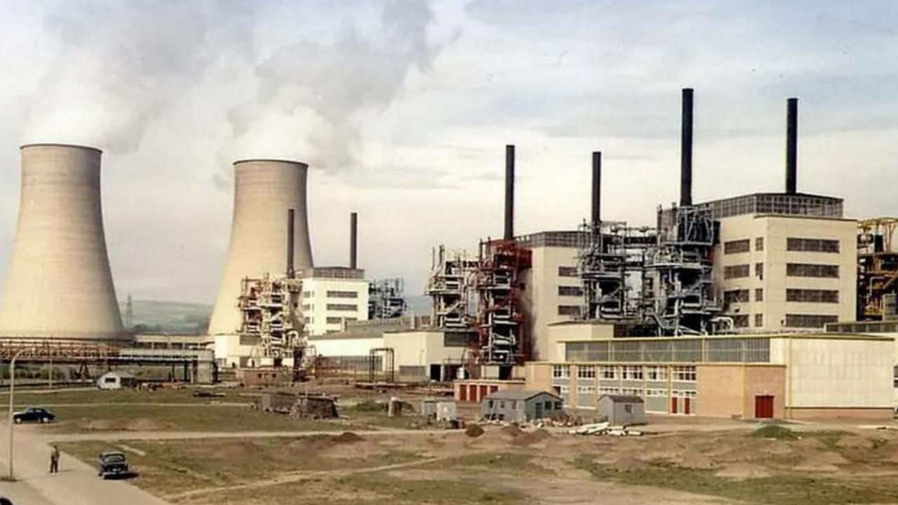 An old photo of Calder Hall. Two towers are emitting white smoke. On the left, are two turbine halls with black chimneys rising out of them.
