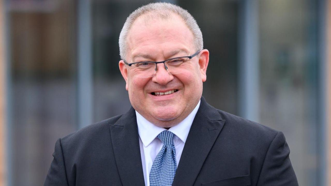 Stephen Drew wearing a black suit jacket, white shirt and blue mottled tie. He wears glasses with a partial black frame and has thinning short grey hair. He is smiling at the camera.