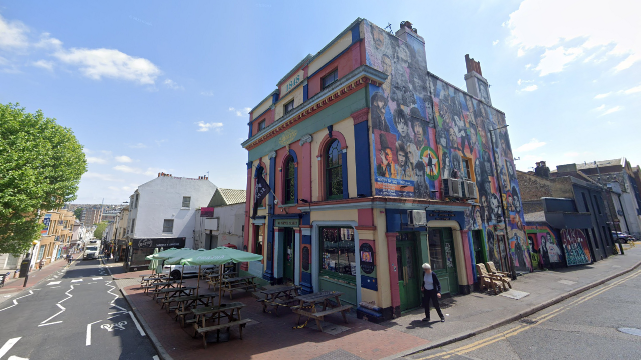 A view from the corner of The Prince Albert pub. The pub is multi-coloured on the front and has paintings of famous musicians' faces on the right side. There are roads running either side. Benches with green umbrellas are outside the front of the pub.