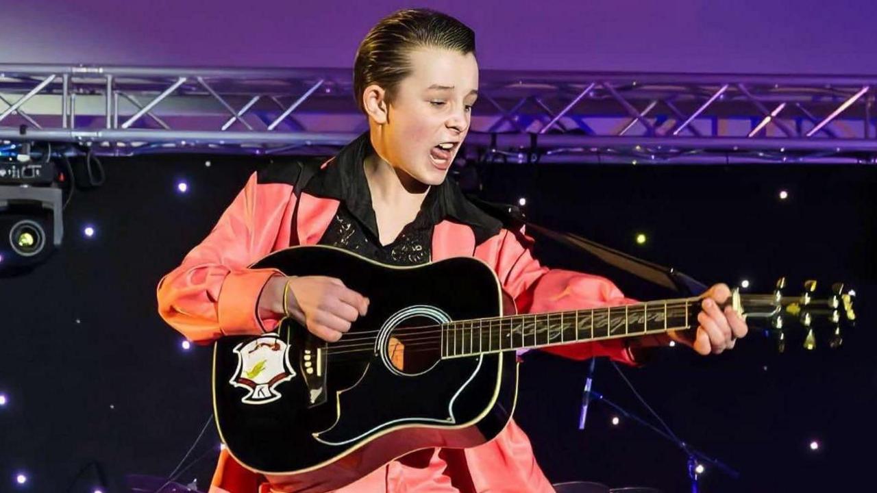 Cooper is on stage playing guitar and singing. He is wearing an orange satin suit and a long lapelled black shirt that has gold sequins on it. His hair is slicked back.