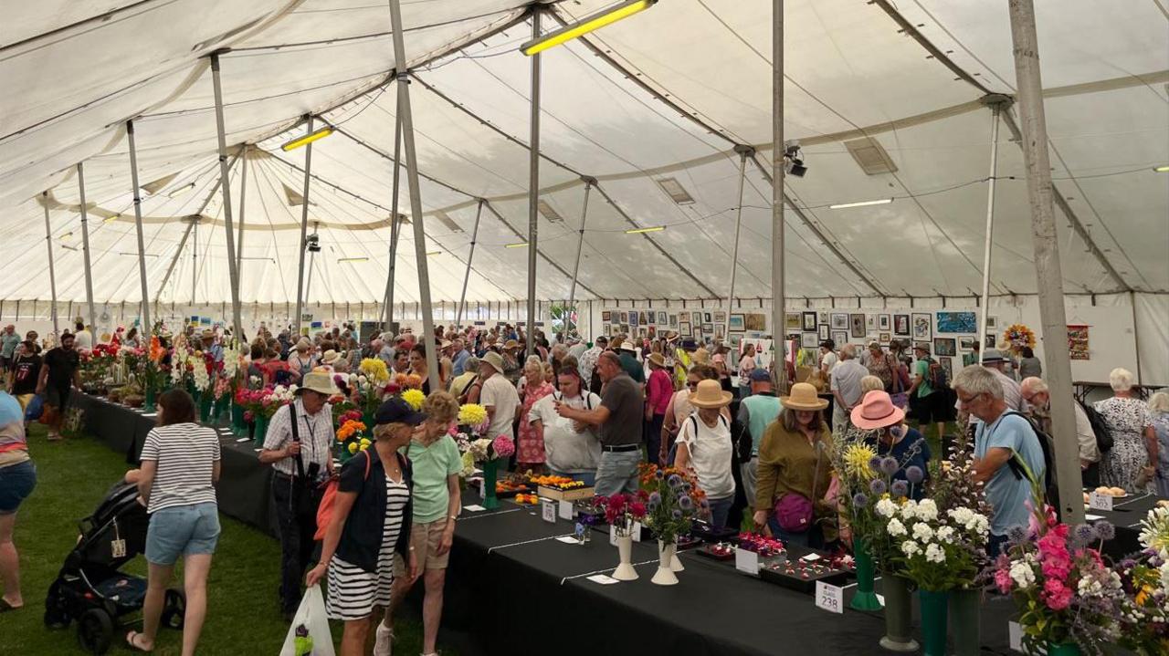 A large crowd browsing inside a marquee. There are long black tables set up with vases and displays of flowers. There are also framed gallery pictures on the walls of the marquee