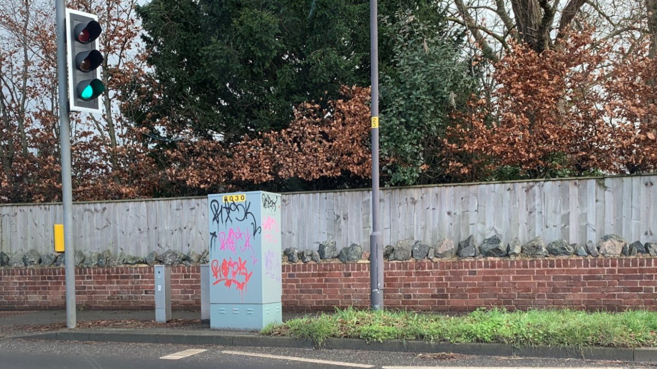 A street with a set of traffic lights and a telegraph pole. An electrical cabinet on the side of the road has been graffitied.