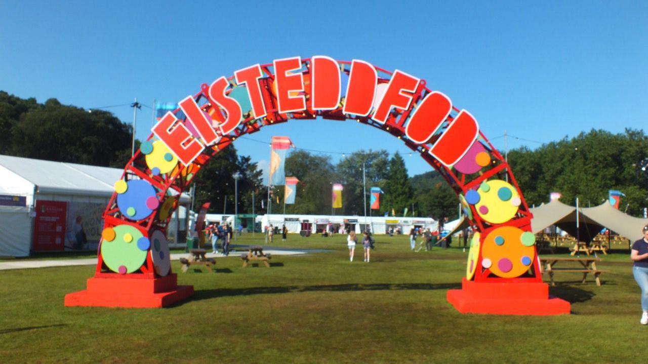 Sign with Eisteddfod spelt out in red over an arch with coloured green, blue, yellow and orange circles festooning the arch. In the background you can see festival flags in blue and yellow and orange and people walking around festival tents