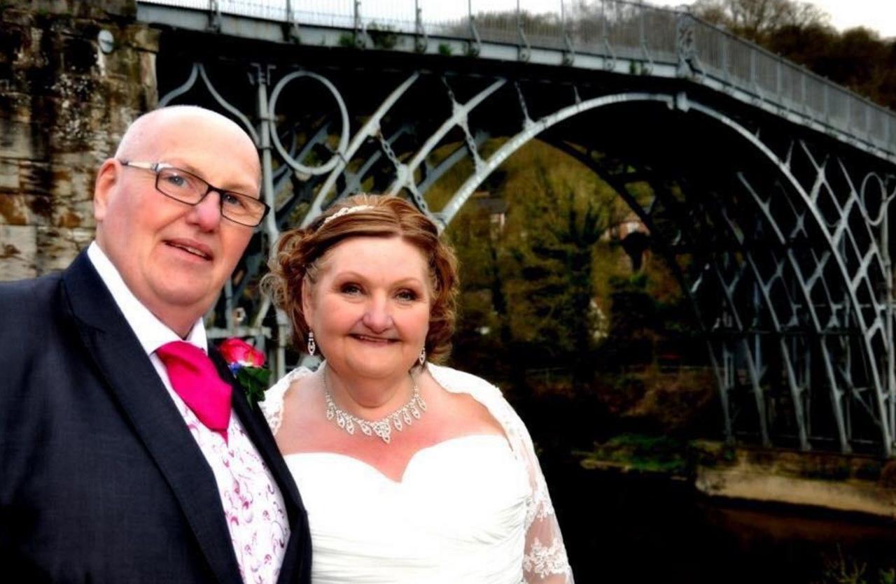 A groom and bride are dressed up near the Iron Bridge in Shropshire. They are on the left of the photo with the man wearing glasses and the woman is in a bridal dress