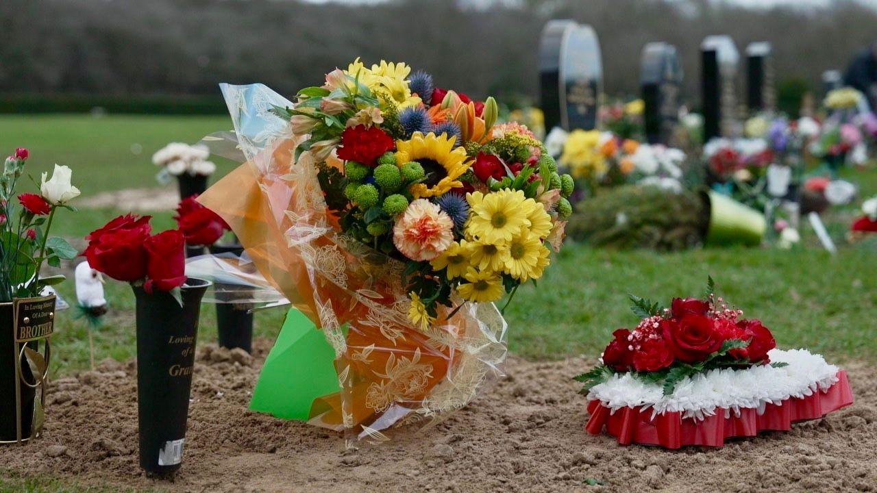 A bouquet of flowers, a wreath and red roses lay on a grave. There are headstones in the background.