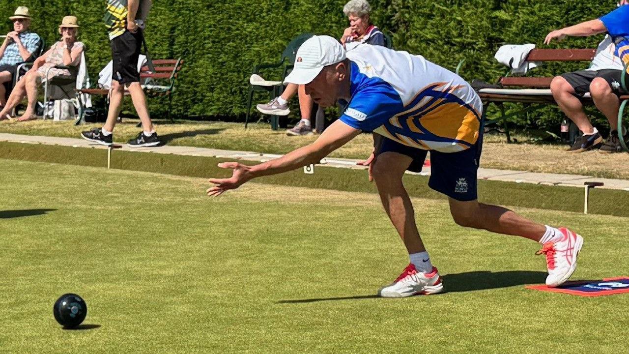 A bowls team-player mid-action as he bowls, with the ball in front of him on the grass. People sit on benches behind in sun hats