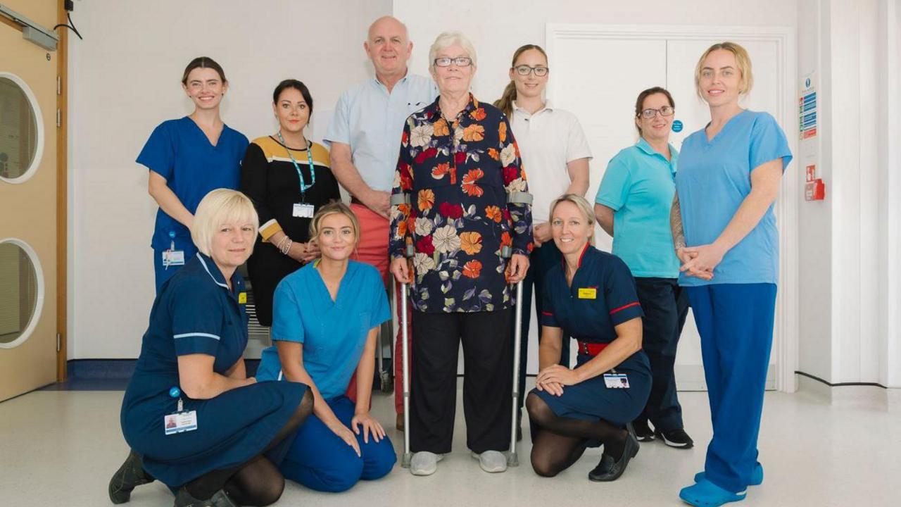 A gathering of men and women in blue medical uniforms around a woman with grey hair, a patterned top and two crutches in a white hospital room