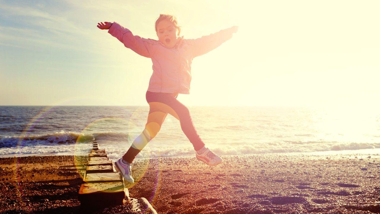 A young girl wearing sports clothes jumps off a board on a beach with her arms raised in the air, the sea is behind and there is a lot of hazy sunshine