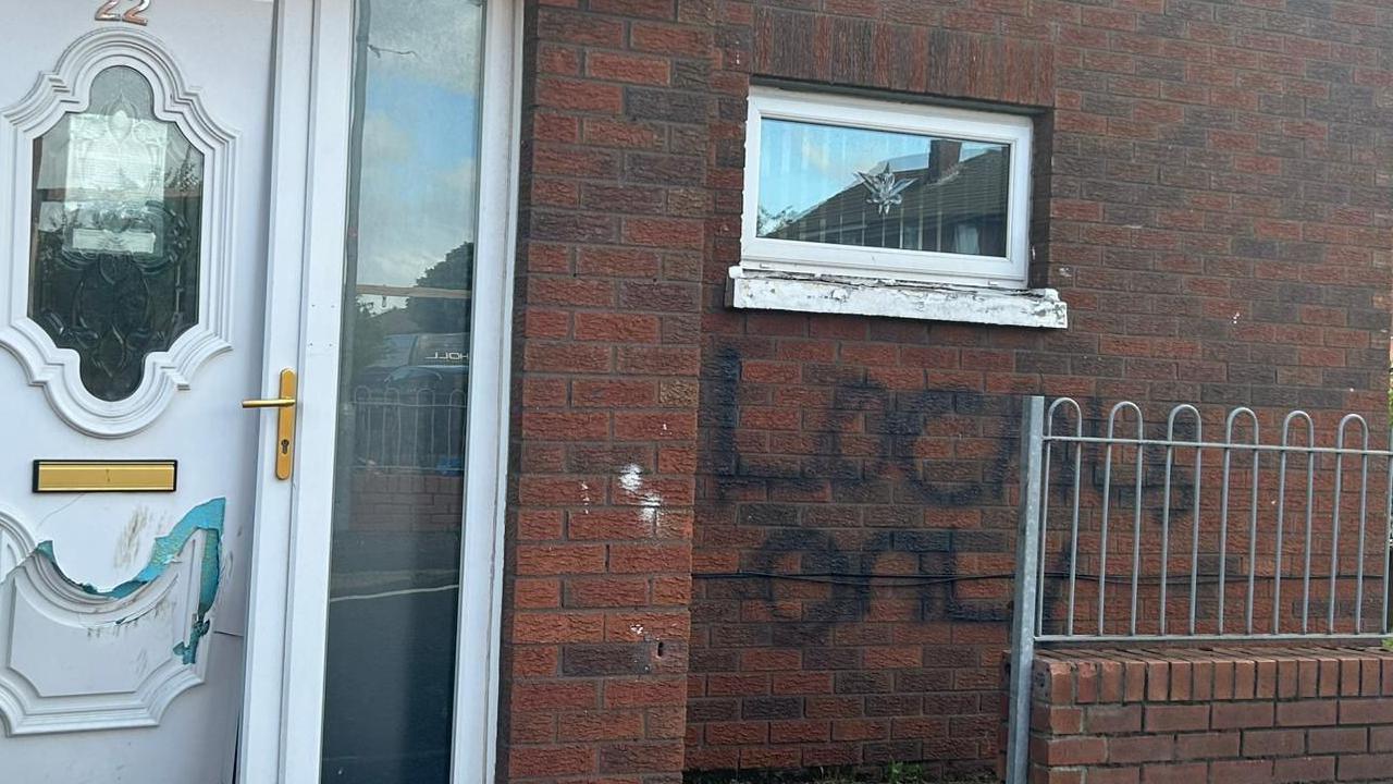 The words "Locals only" are daubed on the side of a house in black paint under a small window. The front door is damaged.