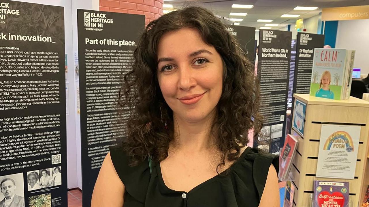 Aynaz Zarif has long brown hair and is wearing a black dress. 
She is standing in a library with books in the background and also standing close to the exhibit. 