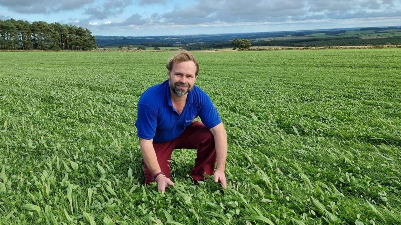 Simon Bainbridge, wearing red trousers and a blue top, is kneeling in a field of pasture with hills behind. 