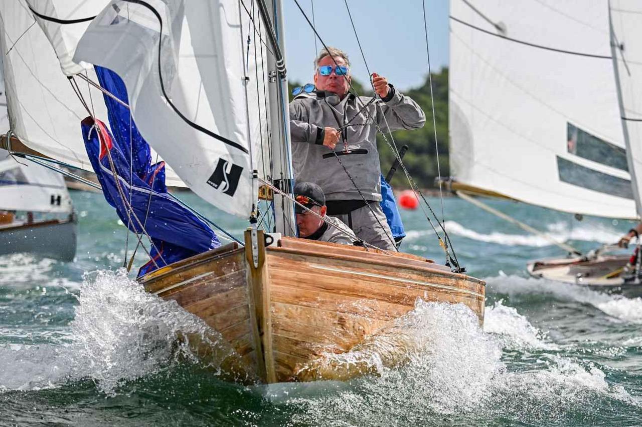 Three sailors on board XOD Xcitation - a varnished wood keel boat with blue and white sails. One of the men is standing and clutching a rope.
