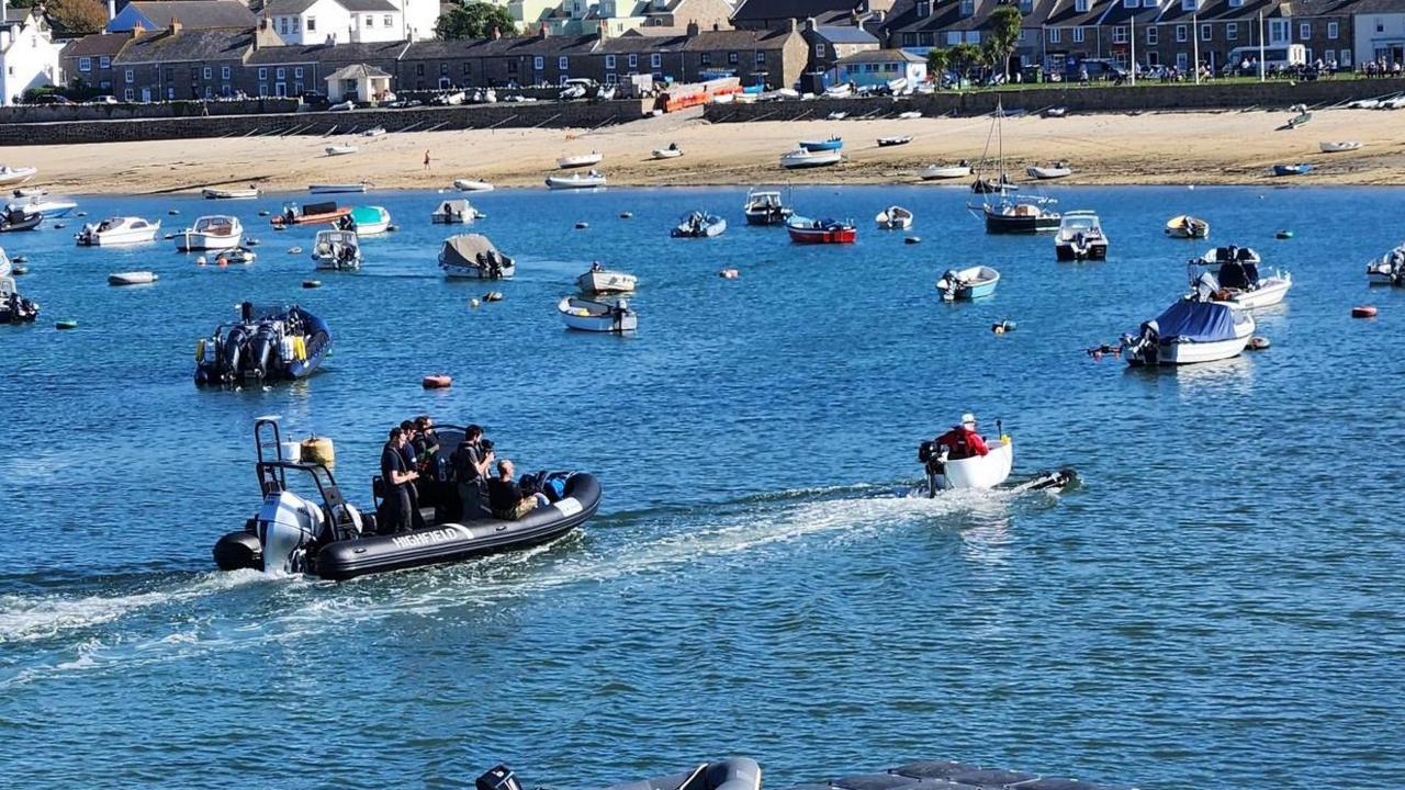 A picture of a number of boats on the water with the beach and houses in the background. There is a boat with a bath tub on it and a support vessel behind it