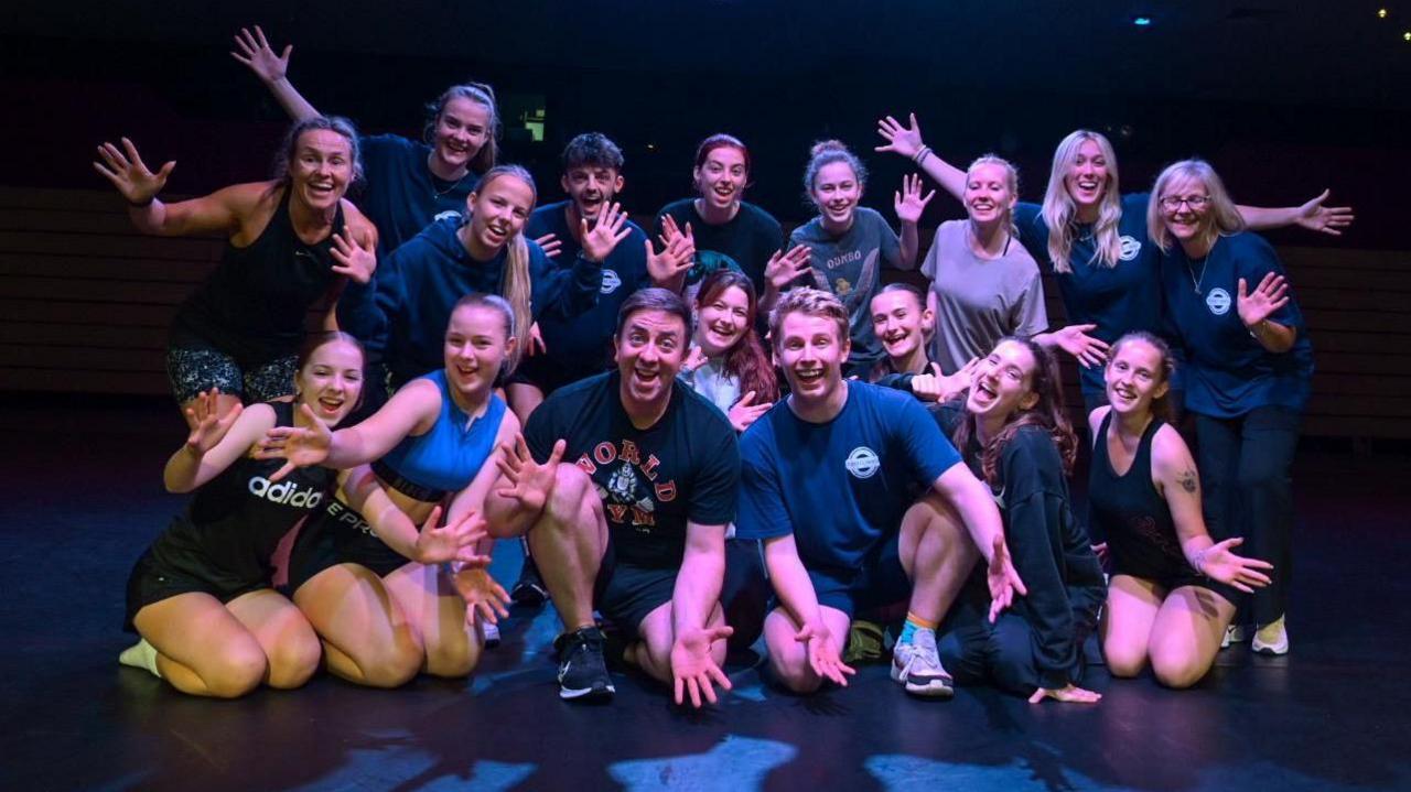 A troop of dancers posing for the camera wearing dark clothes using 'jazz hands'