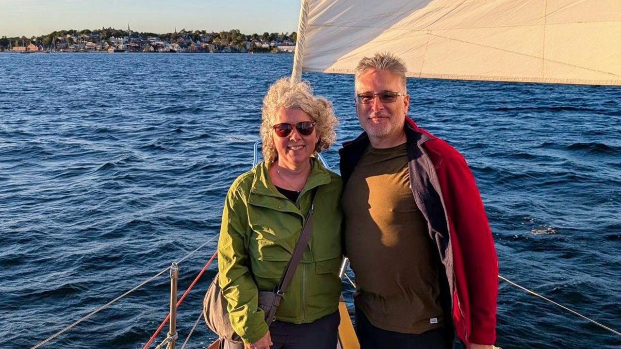 A couple pictured on a boat with blue water behind them and land in the distance. The woman on the right is wearing a green jacket, and the man is wearing a black shirt and a red jacket on top. It is taken during golden hour, with a golden glow from the sun shining on them and the boat.