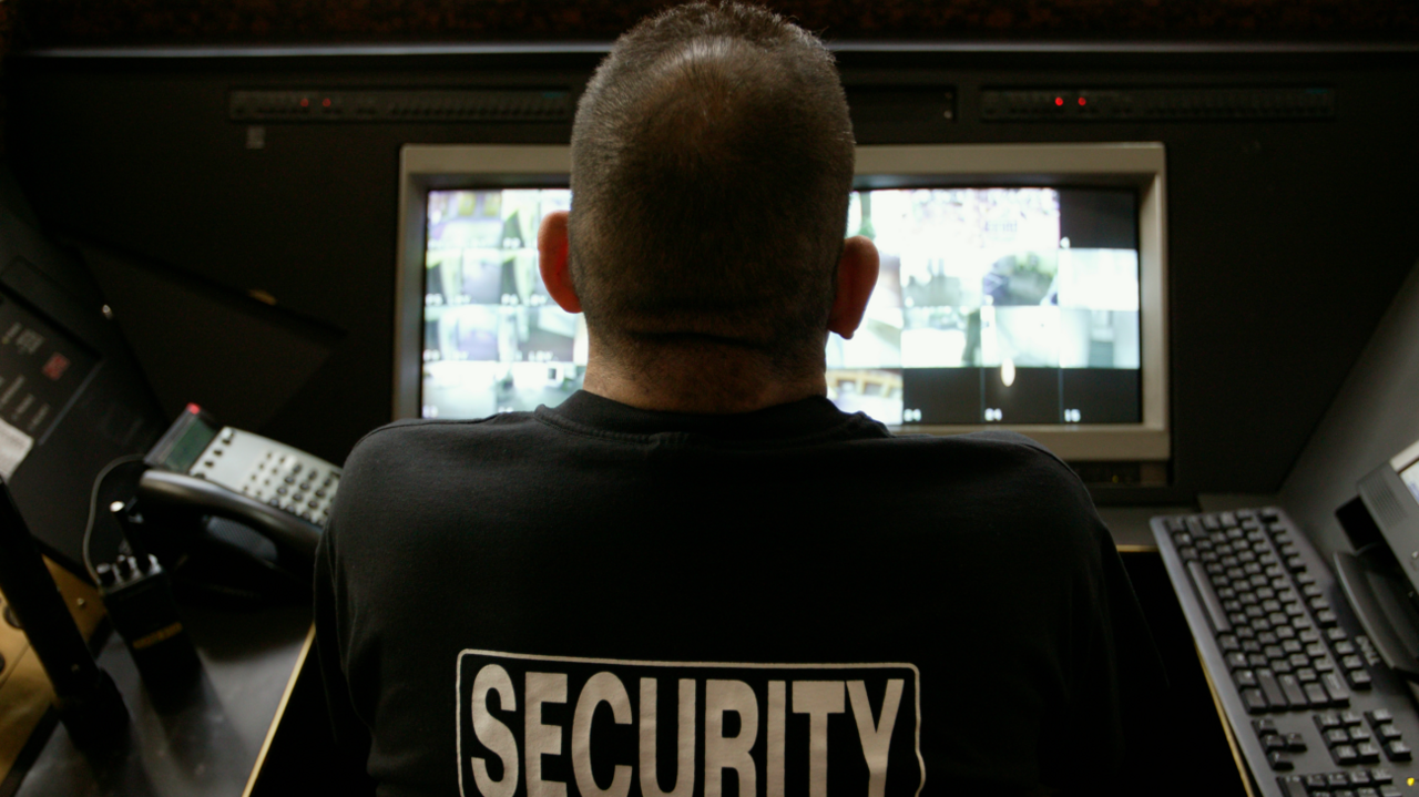 An image of the back of a male security guard wearing a black t-shirt with white writing that reads 'security', sat in front of a screen showing security camera footage