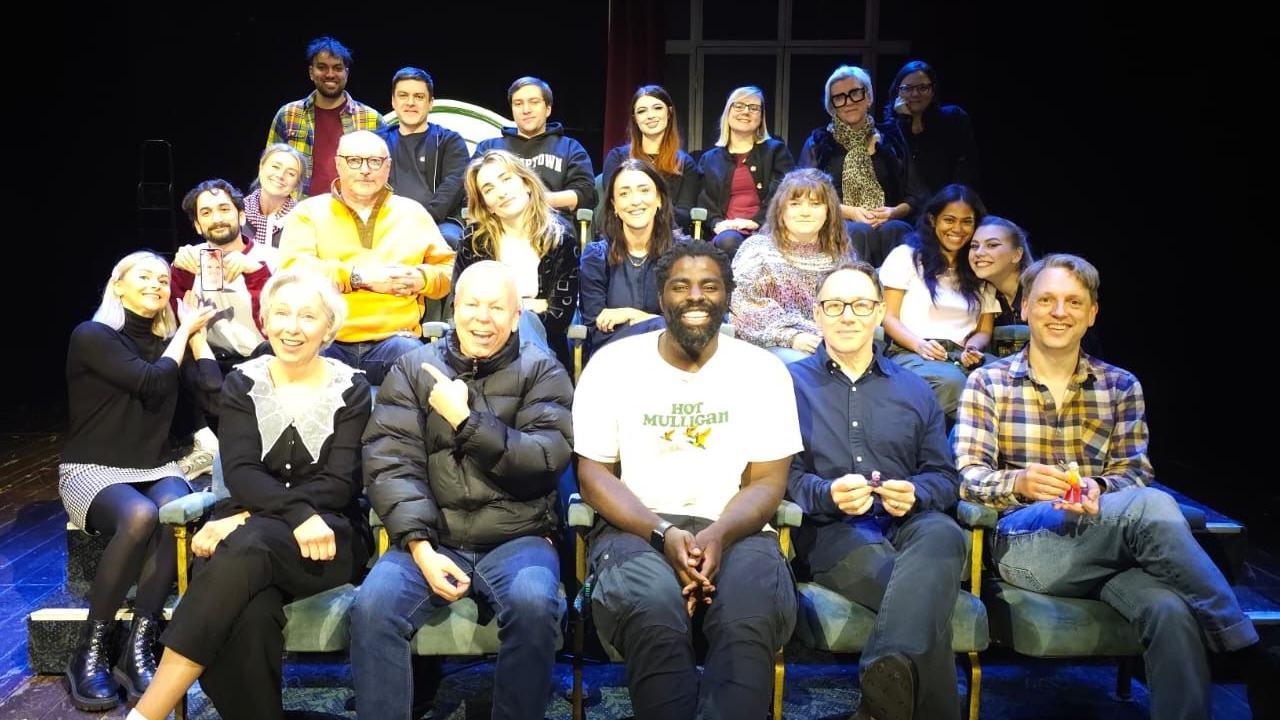 A group of 21 people seated in three rows on a small stage. The seating is arranged in tiers. The lighting is bright on the group, in comparison to the pitch-black background. There is a mixture of males and females on each row, some lean into each other smiling at the camera, others hold small toys in their hand and some point at each other. 