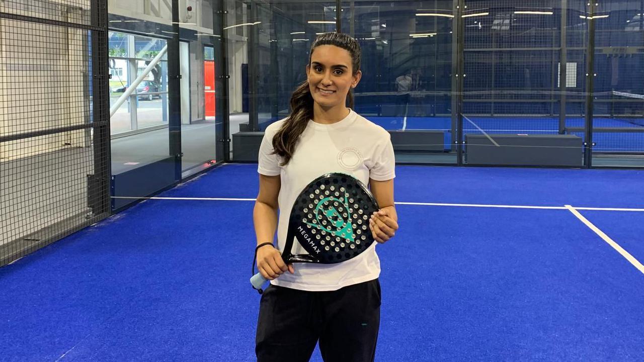 Kiranjit is standing on a blue-floored padel court. The glass walls and black railings of the court can be seen in the background. She is holding a black and blue padel racket in both hands. Kiranjit is wearing a white T-shirt and black leggings. Her long black hair had been tied back into a high ponytail and she is smiling with teeth into the camera.