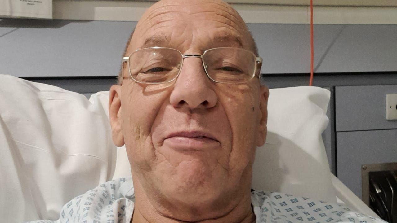 Steven Murfin wears a medical gown and is lying in a hospital bed. He is propped up by a pillow and is wearing glasses as he looks into the camera