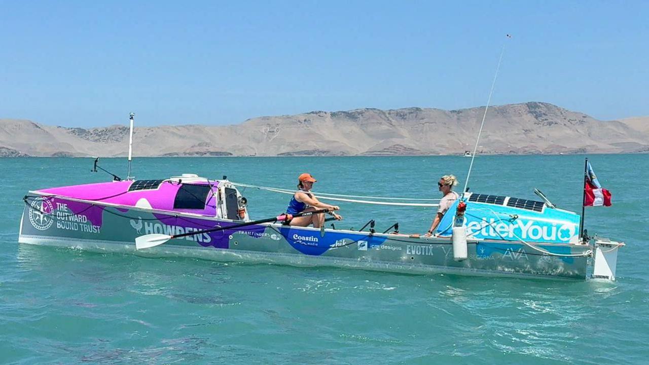 The pair are rowing in the narrow boat in blue sea and barren, dry hills behind them. The sky is blue and clear.