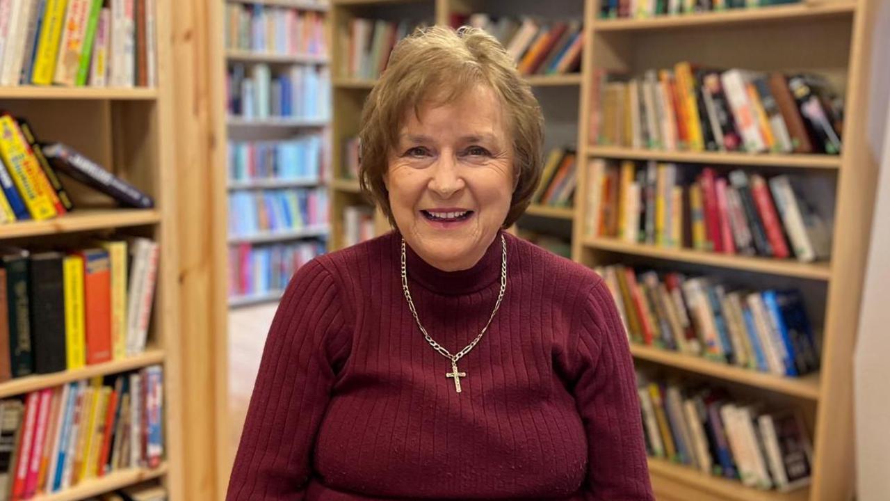 Lady Anne Dodd, wearing a burgundy jumper with a crucifix necklace , smiles at the camera while sitting in a library 