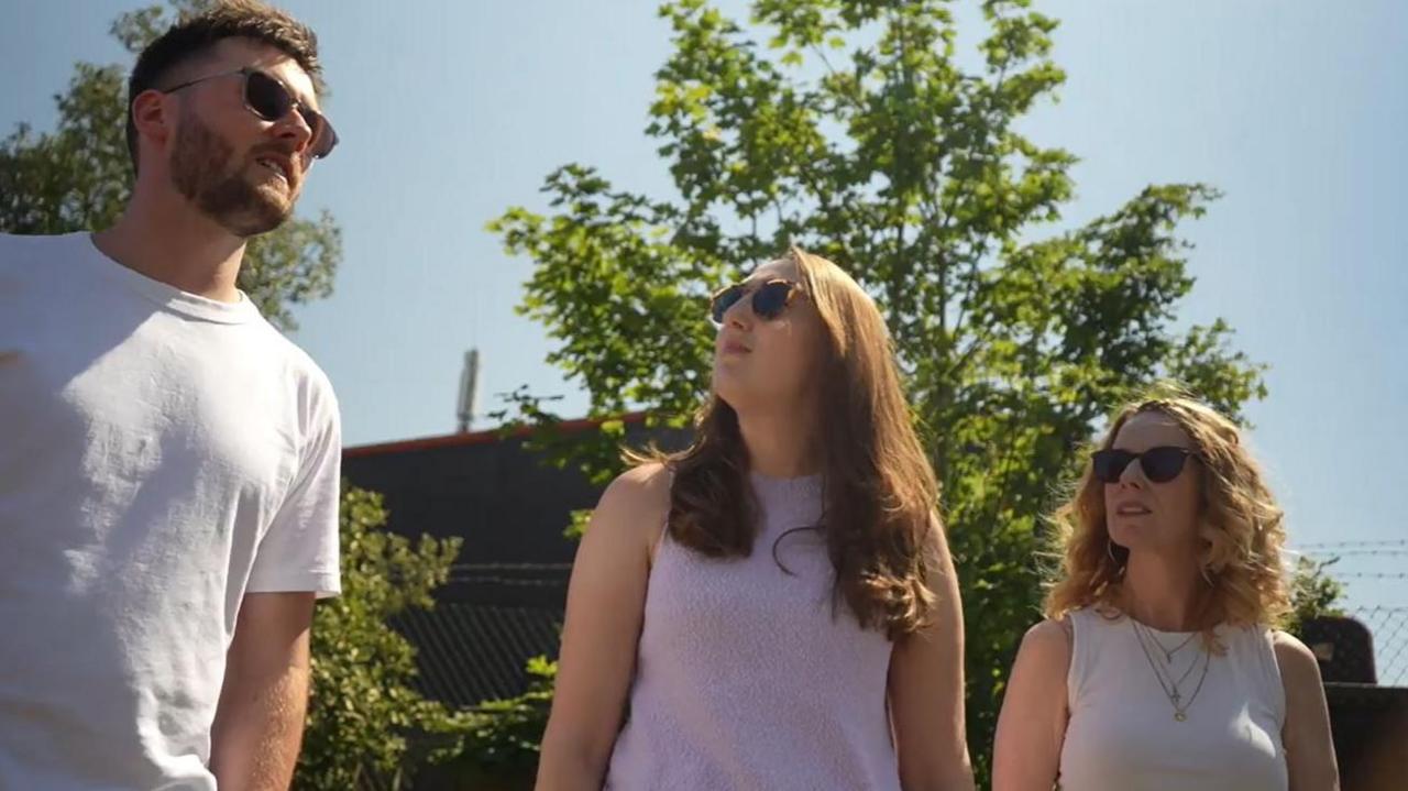 From left to right, Tom, Catherine and Janeane stood next to each other. Each is wearing sunglasses. Tom has cropped brown hair and a beard and is wearing a white t-shirt. Catherine has long brunette hair and is looking at Tom. She is wearing a lilac sleeveless top. Janeane is also looking at Tom. She has wavy blond hair and is wearing a white sleeveless top with necklaces.