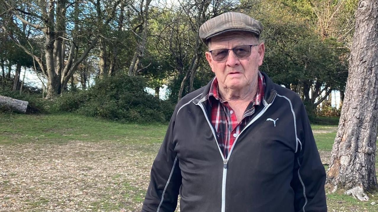 Roly Bessant in a black tracksuit top and flat cap, standing in an area of New Forest woodland