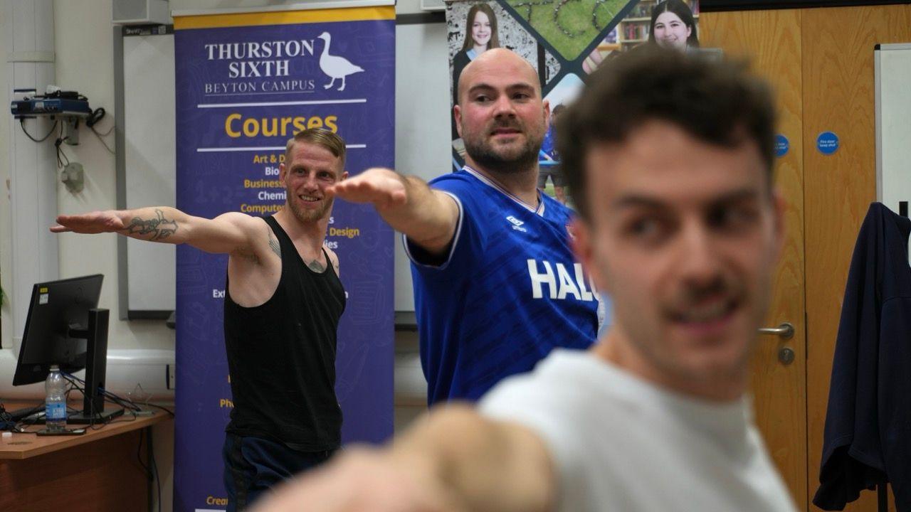 Three members of the group performing the "warrior" pose. Their arms are outstretched as they lunge outwards.
