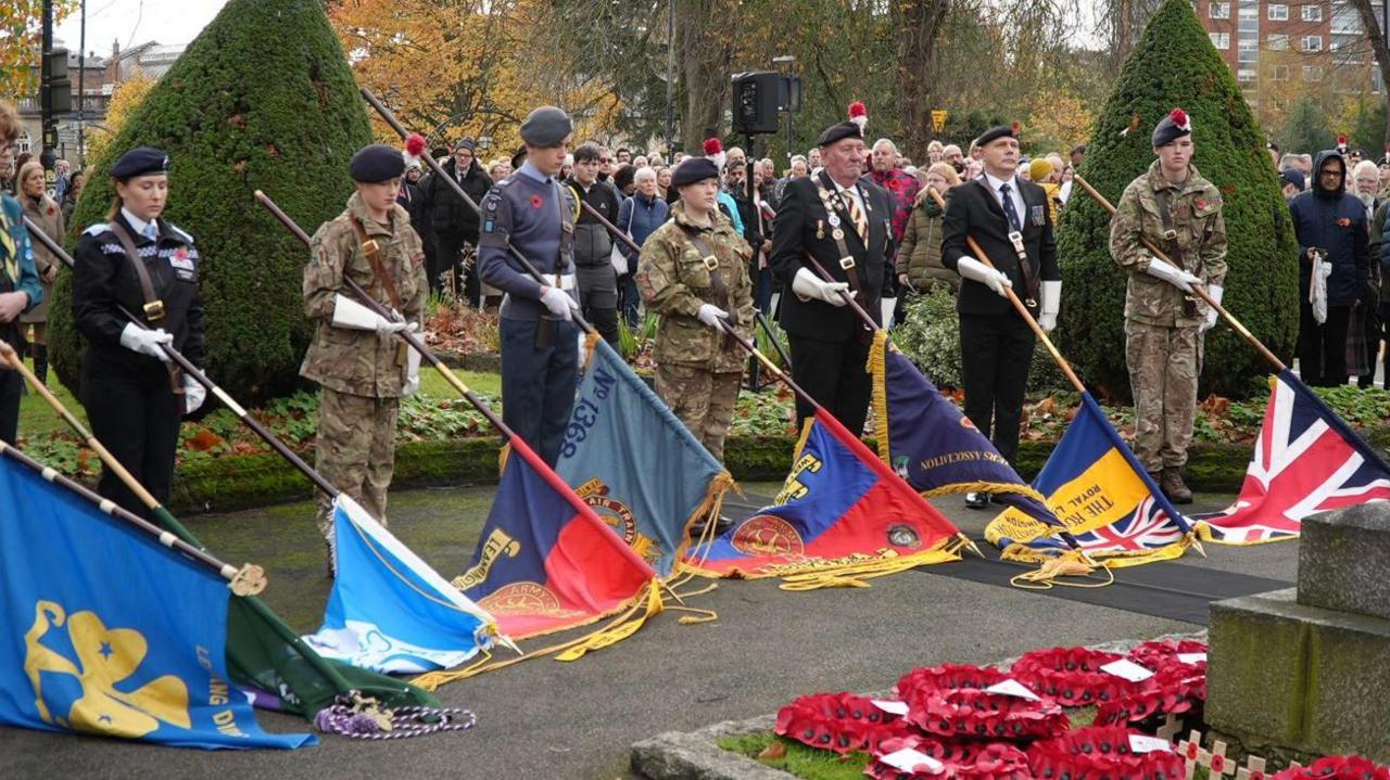 A row of people in military uniform with their heads bowed holding flags in front of them with the ends touching the ground