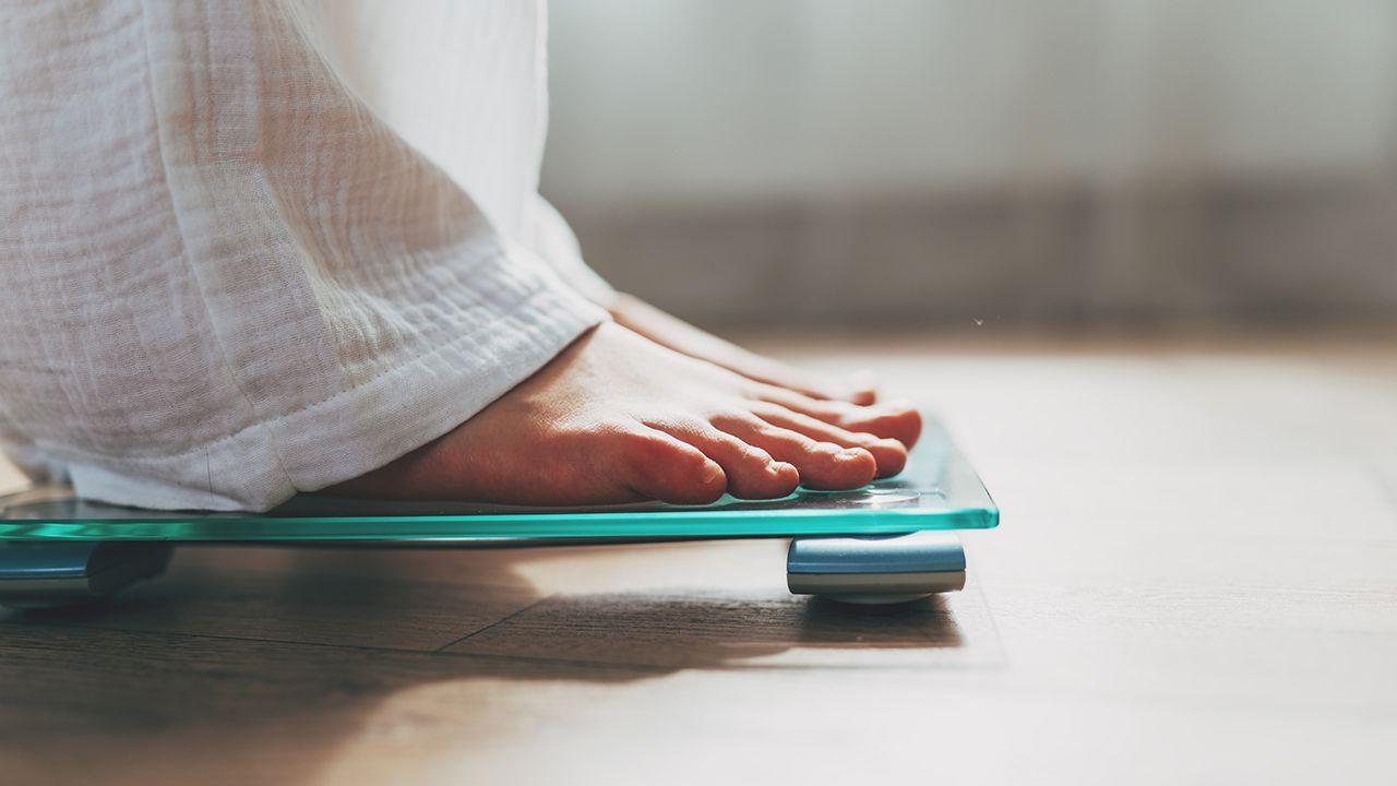 Person weighing themselves on scales. A set of scales can be seen on the floor of a bathroom. The feet of a person is in view as they step on the scales.