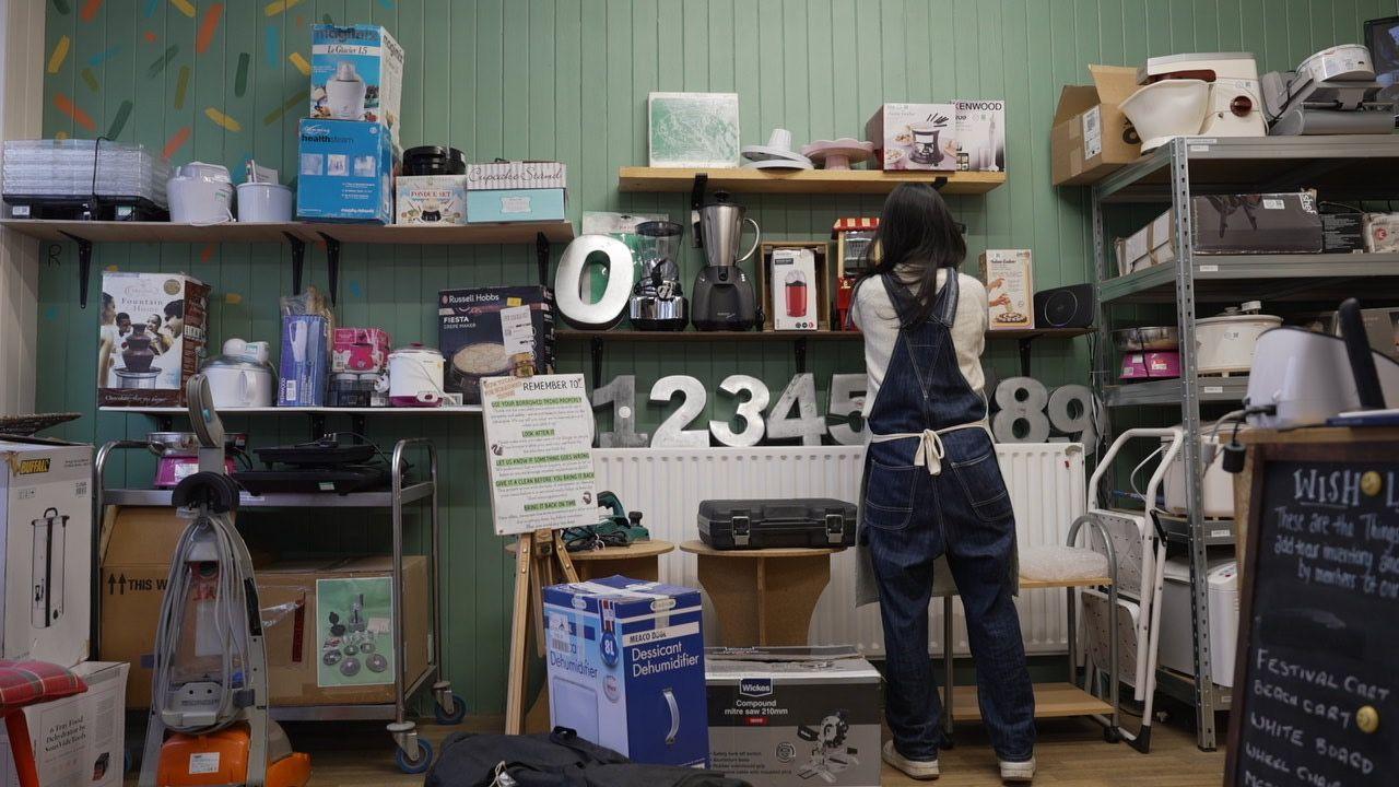 Cecilia wears dark blue dungarees and an apron but has her back to the camera. She is placing kitchen appliances on a shelf in the shed. In the foreground there is a carpet cleaner and a dehumidifier.