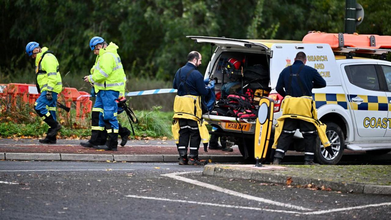 Two men stand at the back of a coastguard rescue vehicle with what appear to be yellow wetsuits rolled down to their waists.  They stand next to the river which as been taped off.