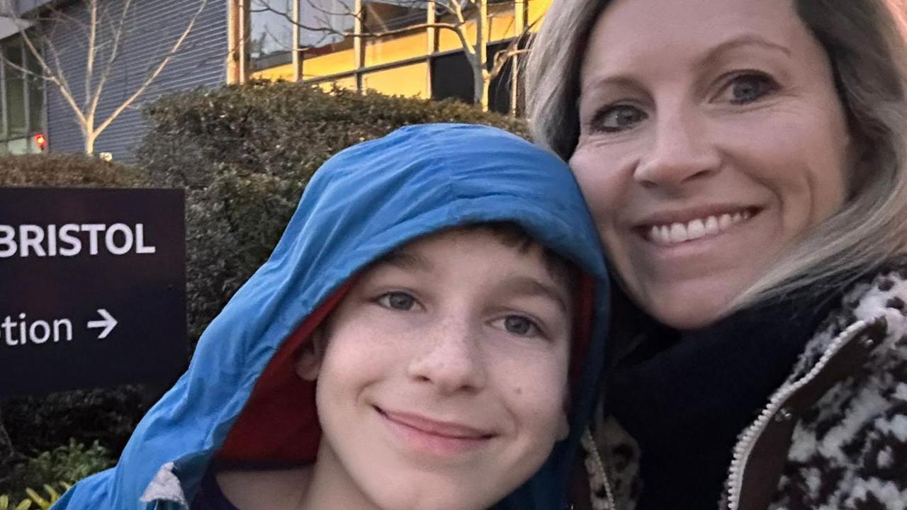 Theo on the left and his mum Katharine on the right. Both are smiling at the camera. Theo has the hood of a blue coat over his head. There is a large building and shrubbery in the background.