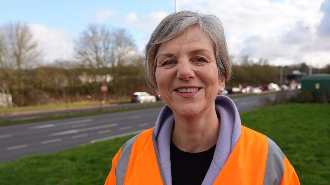 Lilian Greenwood, who has short greying hair with a fringe swept back from her forehead. She is wearing a high-vis orange jacket over a lilac fleece. She is smiling and behind her can be seen grass and beyond it a road and trees.