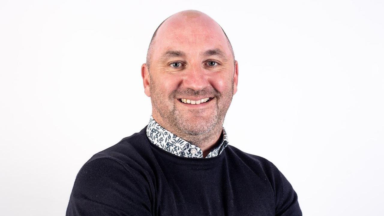 A man with a bald hear wears a black jumper and floral shirt underneath. He is standing against a white background and smiling at the camera.