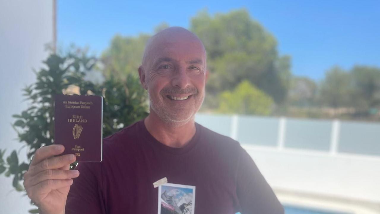 Middle aged man standing outside wearing a burgundy t-shirt holding his Irish passport. He is smiling. The sun is out and the sky is clear blue. 