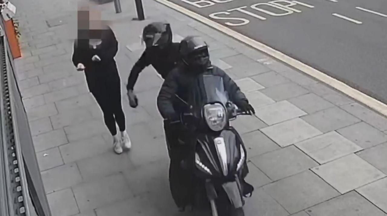 A CCTV image showing a black moped being driven along a pavement by two people wearing all black, snatching a mobile phone from a pedestrian walking along the pavement. One of the people on the moped can be seen holding the phone in their hand moments after snatching it, as the pedestrian recoils.
