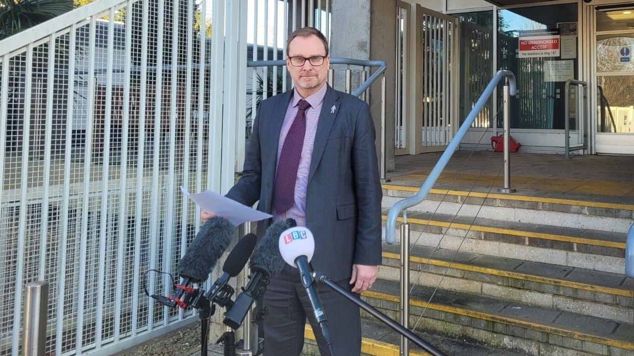 Det Insp Pete Walker standing outside a police building, holding a piece of paper with a statement on it. He is wearing a dark suite, a burgundy tie and a purple shirt, and is looking at the camera. There is a row of microphones, some on a stand and some on poles, held in front of him.