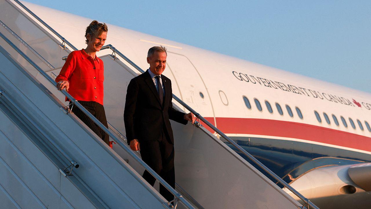 Canada's Prime Minister Mark Carney (right) and his wife Diana Fox Carney disembark an aircraft that says 'Gouvernement Du Canada' along with a red maple leaf, at Paris Charles de Gaulle airport on Monday morning against a clear blue sky. He wears a dark suit and tie and she wears a red shirt and black dress.