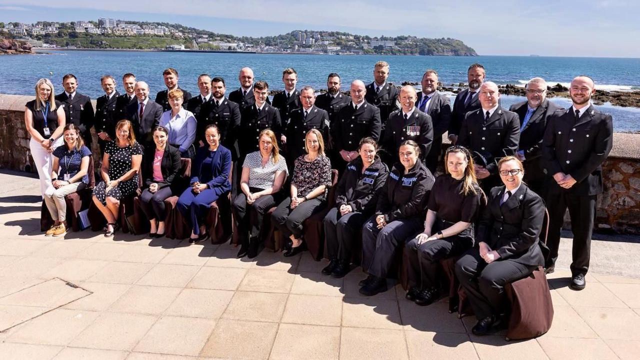 Three rows of police officers in uniform. The front row is sat down on brown chairs and behind them is two rows stood up. There are four officers in uniform on the from right row. The sea is visible behind them. 