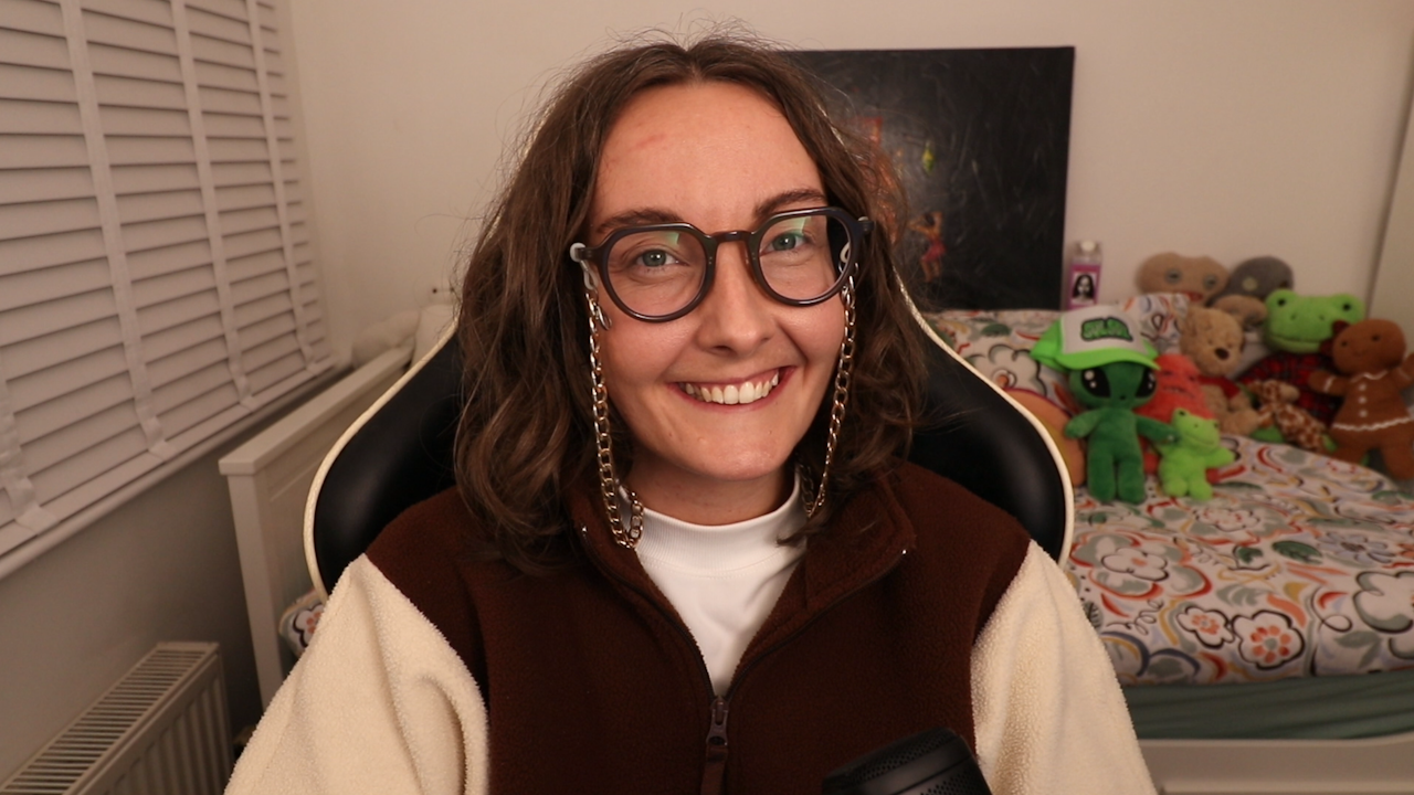 A young woman smiles as she sits in a gaming chair. She wears large glasses with fairly thick black rims and a thick gold chain that allows the spectacles to hang around her neck. Behind her is a day bed with a floral print duvet cover and a collection of soft toys tucked in one corner.