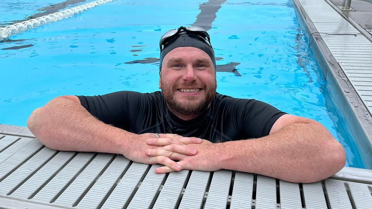 A man with a swim cap and goggles on his head looking and smiling at the camera. He is wearing a dark swimming top while in the water and leaning on the side of the pool