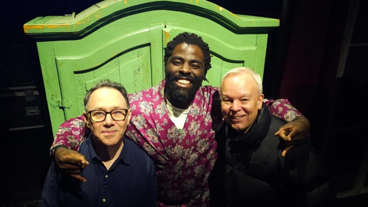 Three men stand in front of a large green double wardrobe. The man on the left has dark hair, wears glasses and has a blue buttoned shirt. The man in the middle is taller then both men, has black braided hair and a black beard, wears a floral dressing gown tied in the middle, and has his arms around the two men with his hands making "rock" symbols. The man on the right has white hair, is smiling at the camera and wears a black puffer jacket.