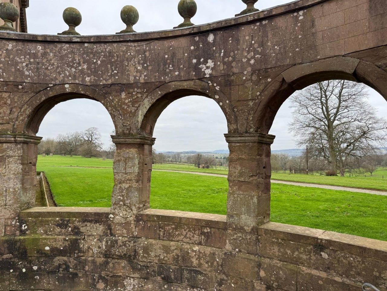 Three arched openings in a curved stone structure with ornate spherical carvings along the top. Parkland and trees are visible through the openings.