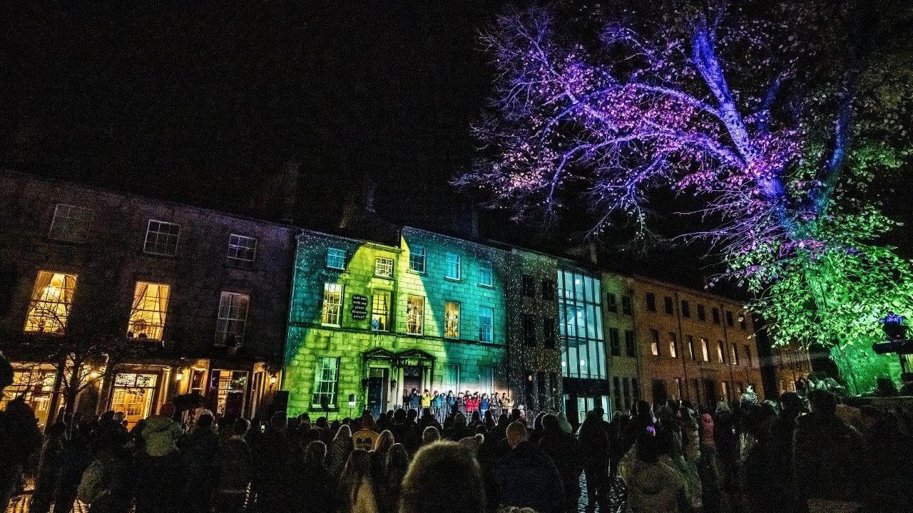 A crowd of people in a Lancaster at night in front of a building lit up with green and blue lights and a tree lit up green and purple